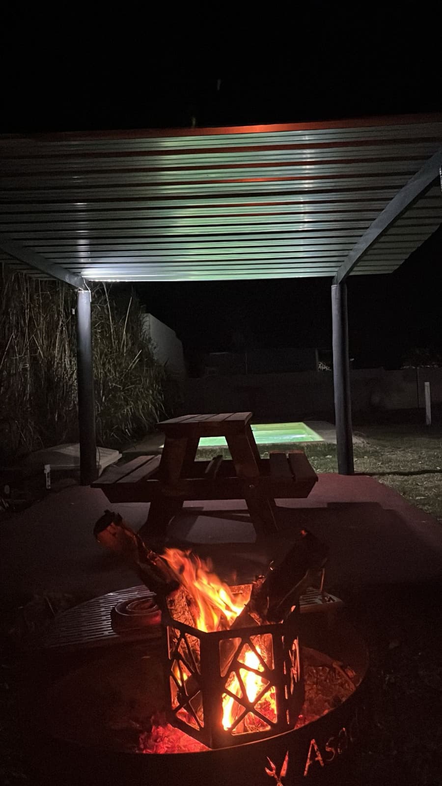 Fogón encendido bajo la pérgola, con la pileta iluminada al fondo